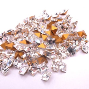 May include: A collection of clear, faceted, square and diamond-shaped rhinestones with gold pyramid-shaped accents. The rhinestones are scattered across a white surface, creating a sparkling effect. Ideal for craft projects.