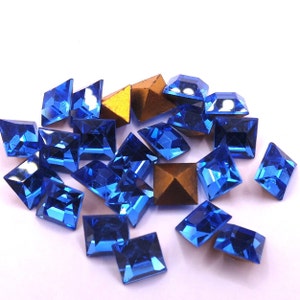 May include: A collection of blue and gold rhinestones in different shapes, including squares, rectangles, and triangles. The rhinestones are scattered on a white surface, reflecting light. Suitable for craft projects and embellishments.