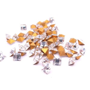 May include: A collection of small, square, clear rhinestones and gold-colored pyramid-shaped embellishments. The rhinestones are faceted, reflecting light, and the gold pieces have a metallic finish. Ideal for craft projects.