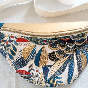 Puede incluir: Una riñonera blanca con una cremallera dorada y una correa azul turquesa. La tela principal presenta un estampado floral colorido con detalles azules, dorados, rojos y negros.