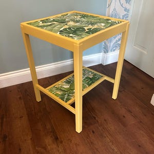 May include: A small, square side table with a yellow painted frame. The tabletop and lower shelf feature a tropical leaf pattern in shades of green and white. The table is approximately 61 cm tall and 46 cm wide.