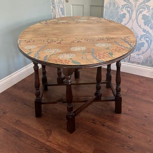 May include: A round wooden drop-leaf table with a natural wood finish top featuring a painted floral design in white, red, and green. The table has dark brown turned legs and a gate-leg base structure that supports the folding side panels.