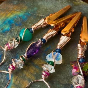 Puede incluir: Cuatro clips de metal en tono cobre con mangos envueltos en alambre y adornados con varias piedras preciosas coloridas, incluyendo cristales verdes, morados y transparentes. Cada clip tiene un bucle circular en la base y pequeñas cuentas.