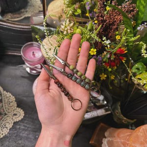 May include: Three metal alligator clip keychains with beaded accents in shades of green, brown, and red. The keychains are held in a hand, with a background of flowers and a lit candle.