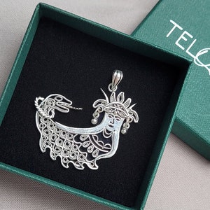 Puede incluir: Un colgante de plata en forma de rostro de mujer con una corona y una serpiente. El colgante está en una caja verde con la palabra "TELOAR" impresa en ella.