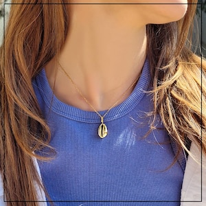 Puede incluir: Collar dorado con colgante de concha. El colgante, con detalles texturizados, cuelga de una fina cadena. El collar se lleva sobre una camiseta azul de canalé.