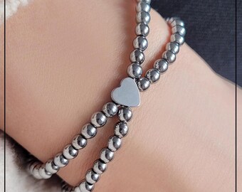 Puede incluir: Un brazalete plateado con un dije en forma de corazón. El brazalete está hecho de pequeñas cuentas redondas.