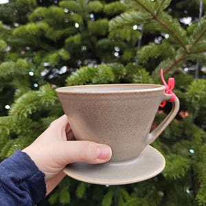 May include: A ceramic coffee dripper in a neutral, earthy tone. The dripper has a conical shape with a handle and sits on a matching saucer. A small red ribbon is tied to the handle. The background is a blurred green Christmas tree.
