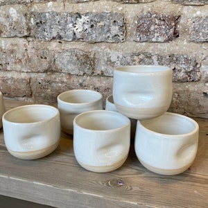 May include: Six white ceramic cups with a distinctive, subtly indented design. The cups have a smooth, glossy finish and an unglazed base. They are displayed on a weathered wooden surface, set against a brick wall.
