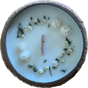 Puede incluir: Una vela blanca con flores secas y una mecha de madera en un cuenco de madera. La vela está rodeada de pequeñas flores blancas y lavanda seca.