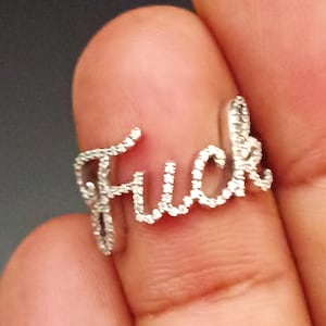 Puede incluir: Un anillo de color plateado que presenta la palabra "Fuck" escrita en una fuente cursiva de estilo manuscrito. La superficie de las letras está incrustada con pequeñas piedras claras y brillantes que captan la luz, creando un aspecto texturizado.