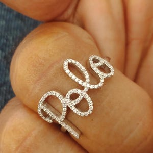 以下が含まれることがあります： 筆記体で「Pele」と書かれたシルバーリング。文字は小さく輝く白い宝石で装飾されています。指にはめられたリング。