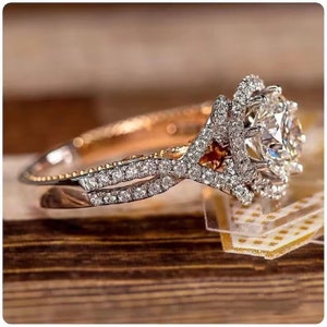 May include: A diamond engagement ring featuring a large, round central stone surrounded by a halo of smaller diamonds. The split band is also embellished with diamonds. The ring rests on a wooden surface.