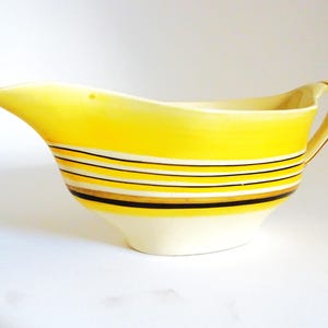 May include: A vintage yellow ceramic gravy boat with a curved spout and handle. The boat features horizontal stripes in black, white, and gold. The base is off-white.