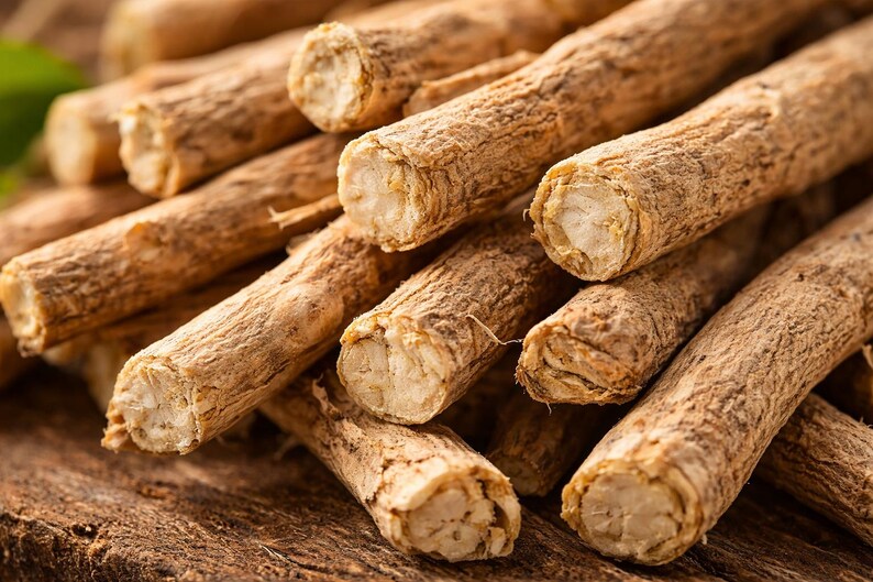 May include: Close-up of a pile of ashwagandha root pieces. The pieces are cylindrical, with a light brown, textured exterior and a lighter, fibrous interior. The roots are stacked on a dark wooden surface, showcasing their natural appearance.