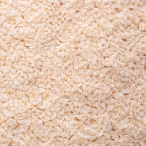May include: Close-up of a pile of light beige psyllium husk, a dietary fiber supplement. The texture is granular and slightly coarse, with small, irregular flakes and particles. The color is a natural, off-white hue.