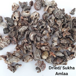 Könnte beinhalten: Nahaufnahme von getrockneten Amla-Früchten, auch bekannt als indische Stachelbeere. Die Fruchtstücke sind eine Mischung aus Braun und Beige mit einer runzligen Textur. Der Text "Dried/ Sukha Amlaa" ist sichtbar.