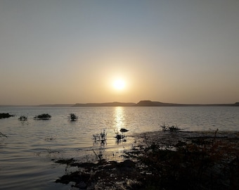 May include: A serene landscape photograph of a lake at sunset. The sun, a bright orb, reflects on the water's surface, creating a shimmering path. The sky transitions from a light blue to a warm golden hue, with silhouettes of distant hills.