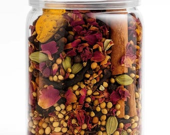 May include: A clear glass jar filled with a blend of spices and dried botanicals. The contents include rose petals, cardamom pods, cinnamon sticks, and small, round seeds. The jar has a silver lid and is set against a white background.