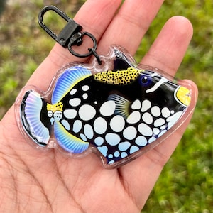 May include: A black and white triggerfish keychain with blue and yellow accents. The fish is made of resin and has a clear plastic backing. The keychain is attached to a black metal ring.