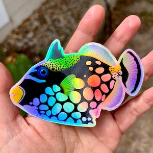 May include: A holographic sticker of a black and rainbow colored fish with white spots. The fish has a green and yellow fin and a purple tail.
