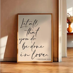 May include: A white framed print with black calligraphy that reads "Let all that you do be done in love. 1 Corinthians 16:14". The print is hanging on a wall in a room with a wooden floor.