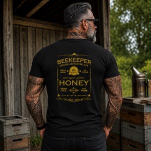 May include: Black t-shirt with a gold graphic that reads "BEEKEEPER" and "HONEY". The design includes text such as "Genuine Beekeeping Age", "Best Quality", "100% Pure", and "Local Raw Unfiltered". The shirt is worn by a person with tattoos.