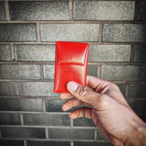 May include: A small, red leather wallet or case. The wallet is held in a hand against a brick wall background.