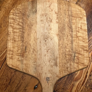May include: A light-colored wooden pizza peel with a wide, paddle-shaped head and a long handle. The wood grain is prominent, with a natural, textured appearance. A small, etched design is visible on the handle. The peel rests on a wooden surface.