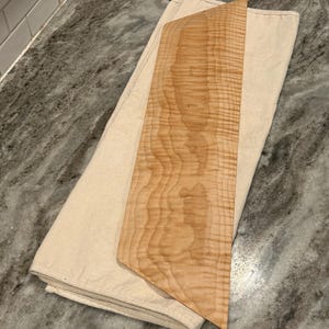 May include: A rectangular wooden board with a light tan color and a visible wavy grain pattern, placed on top of a folded, off-white canvas cloth. The board features straight edges and angled corners, showcasing the natural texture of the wood grain.