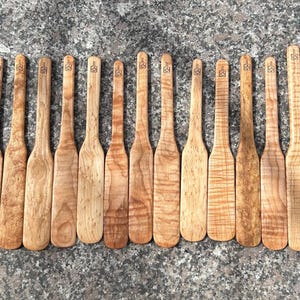 May include: A collection of thirteen light brown wooden spatulas arranged in a row. Each spatula features a long handle and a flat, wide blade with a small, square-shaped logo stamped near the top of the handle, showcasing natural wood grain patterns.