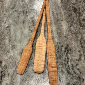 May include: Three light brown wooden spatulas with a wavy grain pattern. Each utensil features a long, slender handle and a flat, rounded blade. One spatula is marked with a small square logo containing a maple leaf and a bottle silhouette.