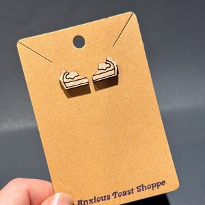 May include: A pair of wooden earrings shaped like pie slices. The earrings are on a brown card with the text "Anxious Toast Shoppe".