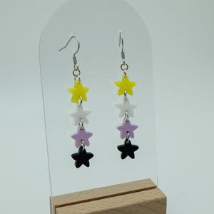 May include: A pair of dangle earrings with four stars on each earring. The stars are yellow, white, lavender, and black.