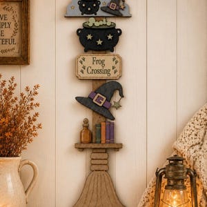May include: A vertical wooden wall hanging featuring a stacked design with a cloud, a black cauldron with green bubbles, a sign reading Frog Crossing, a black witch hat, a small shelf with a bottle and books, and a broomstick base.