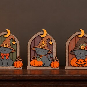 May include: Three arched wooden decorations featuring grey cats wearing pointed witch hats. Each cat is positioned within a frame with a crescent moon and star cutout. The cats are accompanied by orange pumpkins, with one cat sitting inside a carved pumpkin.