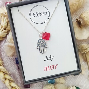 May include: A silver necklace with a hamsa hand charm and a red gemstone pendant. The necklace is in a box with the text "July RUBY" on it.