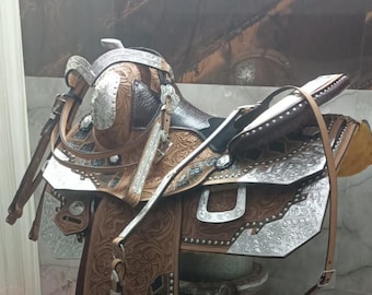 May include: A Western-style horse saddle with detailed silver and brown leather. The saddle has decorative tooling, stirrups, and a high cantle. It is displayed on a small wooden stool, highlighting its craftsmanship.