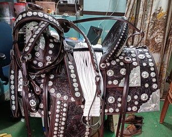 Puede incluir: Una silla de montar de cuero marrón oscuro detallada con intrincados grabados florales y detalles plateados. La silla presenta numerosos adornos brillantes y un cabezal a juego, mostrando la artesanía y el diseño ecuestre.