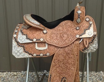 May include: A brown leather saddle with silver accents and intricate tooling. The saddle is on a metal stand and has a black seat.