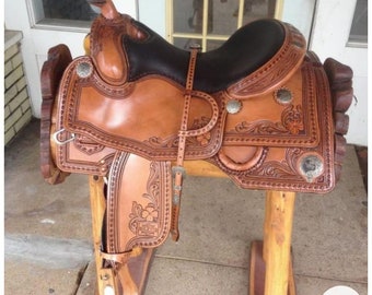 May include: A brown leather saddle with intricate floral tooling and silver accents. The saddle is mounted on a wooden stand.