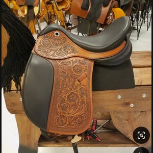 Puede incluir: Una silla de montar de cuero marrón con intrincados detalles florales. La silla de montar es negra en el asiento y tiene una solapa de cuero negro. La silla de montar se muestra en un soporte de madera.