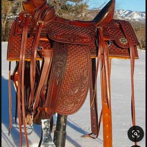 May include: A brown leather saddle with intricate stitching and tooling. The saddle is mounted on a metal stand and is set against a snowy background.