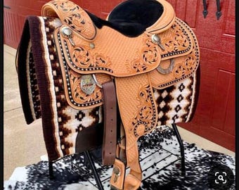 May include: A brown leather saddle with intricate tooling and a black seat. The saddle is on a black metal stand and a cowhide rug.