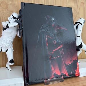 Puede incluir: Un libro con una cubierta oscura que presenta una figura sentada con un abrigo largo, con un pájaro en el hombro y un fondo rojo. El libro está sostenido por un soporte transparente, flanqueado por dos figuras blancas de Stormtroopers.