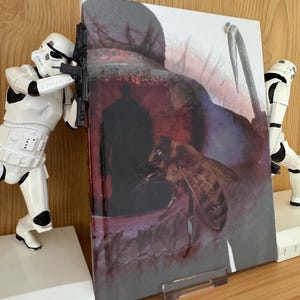 Puede incluir: Un libro con una abeja y una hoz en la portada, sostenido por dos sujetalibros blancos con forma de Stormtrooper. La portada del libro tiene una combinación de colores oscuros con una abeja y una hoz.