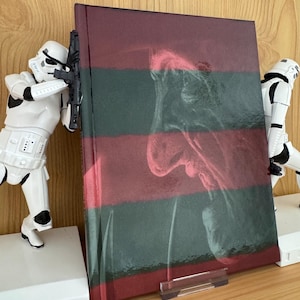 Puede incluir: Un libro de tapa dura con un diseño a rayas rojas y verdes, con una imagen borrosa de una cara. Dos figuras blancas de Stormtroopers están a ambos lados del libro, como si lo protegieran.