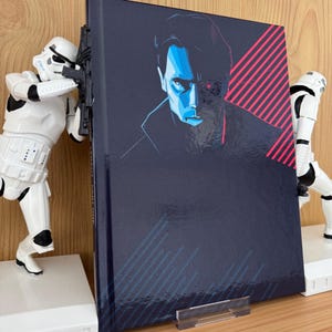 Puede incluir: Un libro de tapa dura con una cubierta azul oscuro que presenta un retrato estilizado de un hombre con acentos azules y rojos. El libro está apoyado por dos figuras blancas de Stormtroopers. El fondo es una superficie de madera marrón claro.