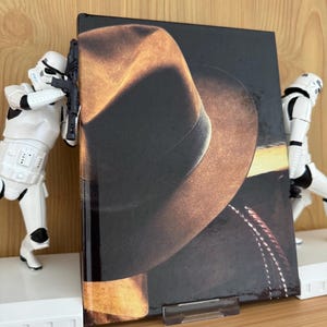 Puede incluir: Un libro de tapa dura con una imagen grande de un sombrero de vaquero en la portada, sostenido por dos figuras blancas de Stormtroopers. El sombrero es marrón y negro, con un ala y una copa detalladas. El libro está sobre una superficie de madera.