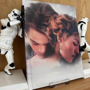 Puede incluir: Un libro con una portada de cartel de película, sostenido entre dos sujetalibros blancos de Stormtrooper. La portada del libro presenta una imagen romántica de un hombre y una mujer. Los sujetalibros tienen forma de Stormtroopers de Star Wars.